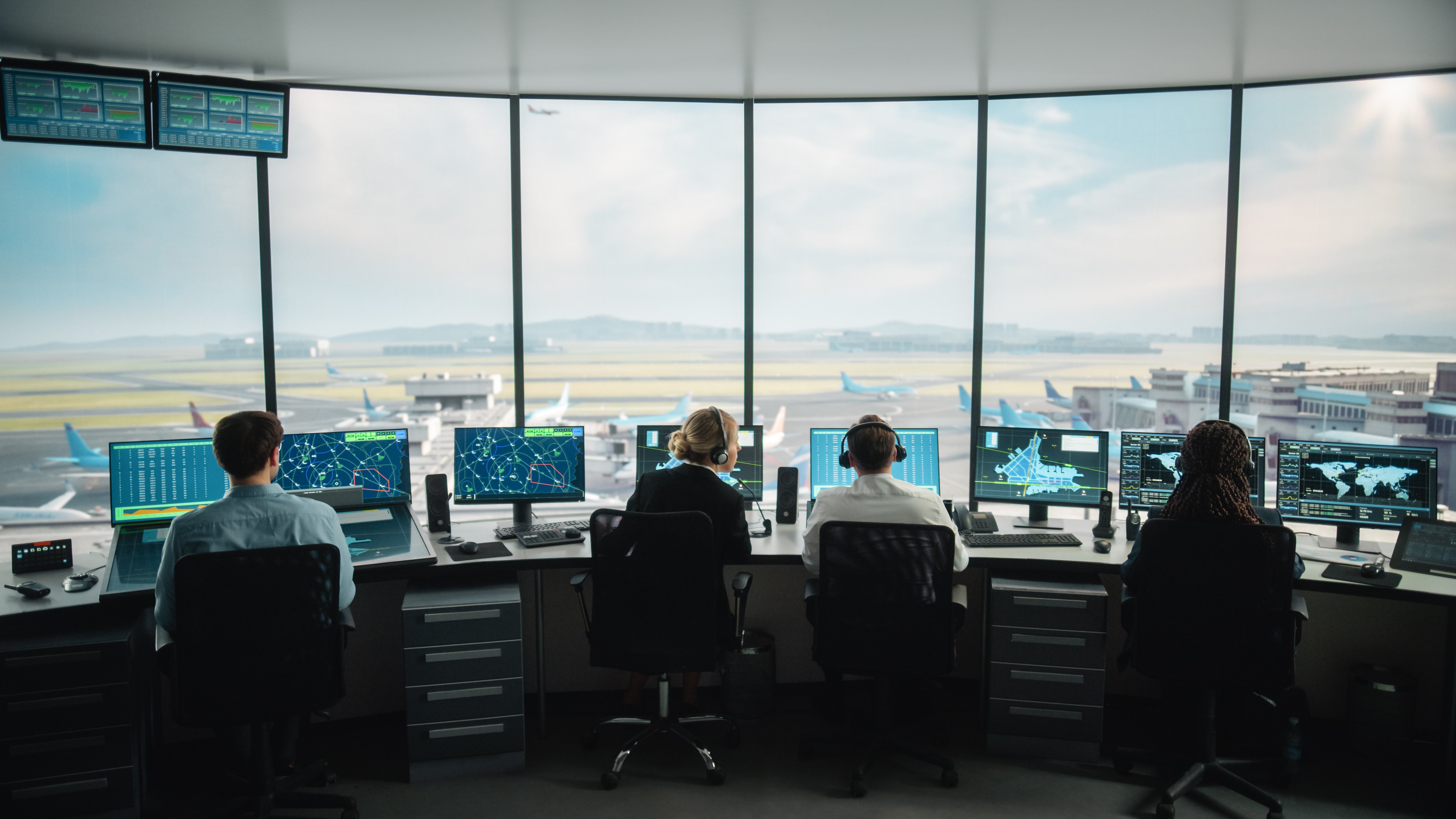 Four air traffic controllers are seated at workstations in an airport control tower, each monitoring multiple screens displaying radar, flight paths, airport maps, and global data. Large panoramic windows offer a clear view of the airport runway, taxiing airplanes, and terminal buildings. The team coordinates aircraft movements in a high-tech, secure environment