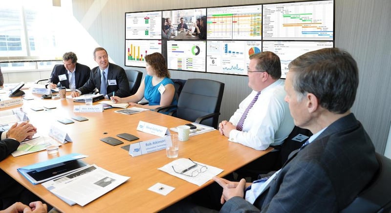 5 employees have a discussion while sitting at a table in a meeting room with a video wall displaying data visualizations