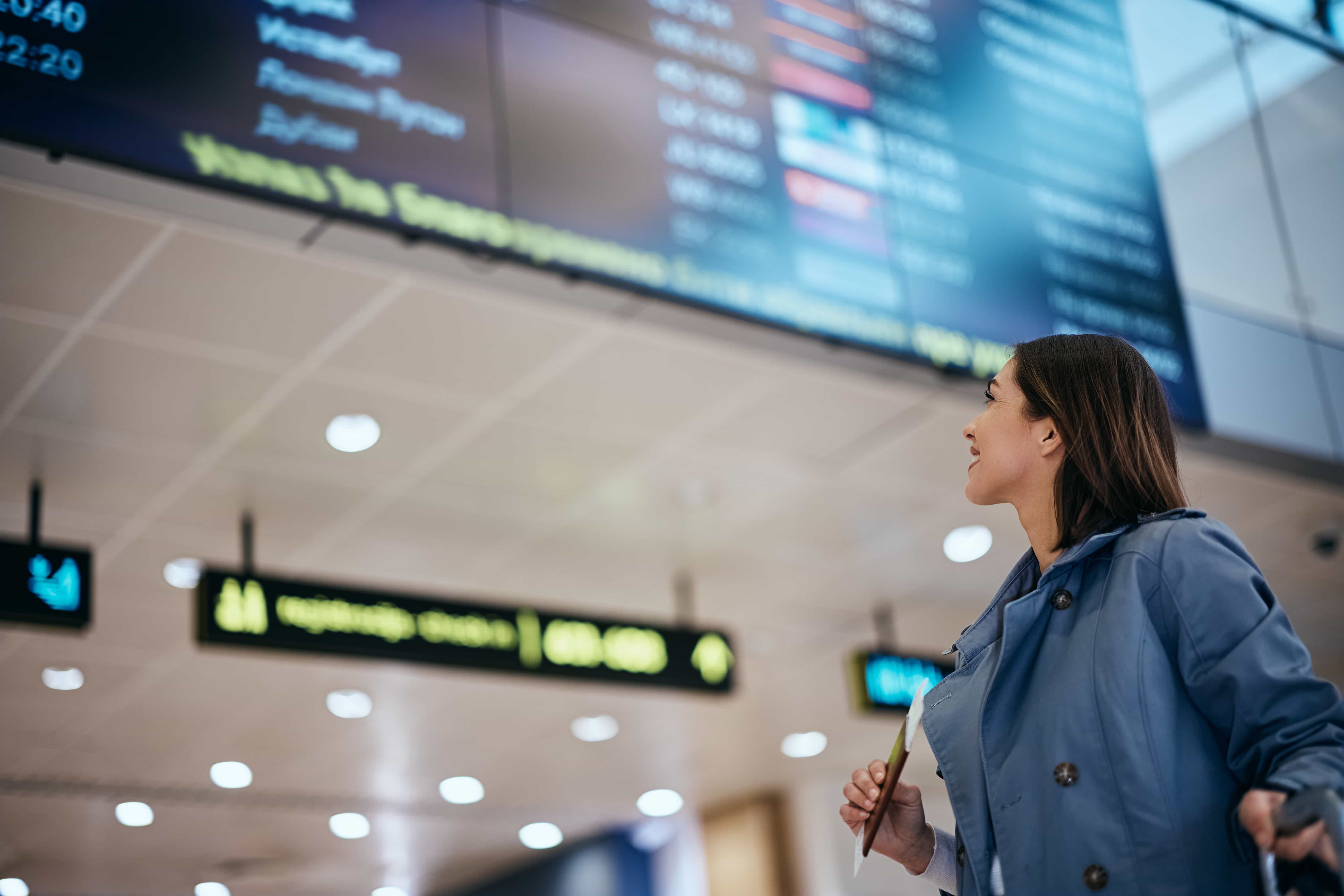  A woman in a light blue jacket and white shirt looks up at a large departures board in an airport. She is smiling slightly and holding what appears to be a passport or ticket in her right hand. The departures board, which is out of focus, displays various flight information. The airport ceiling with lights is also visible in the background.