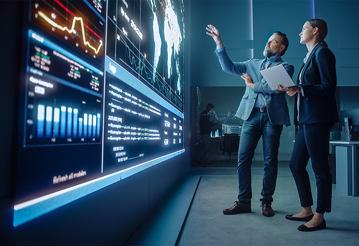 Man and woman in an office having a discussion while referring to a large video wall displaying data dashboards