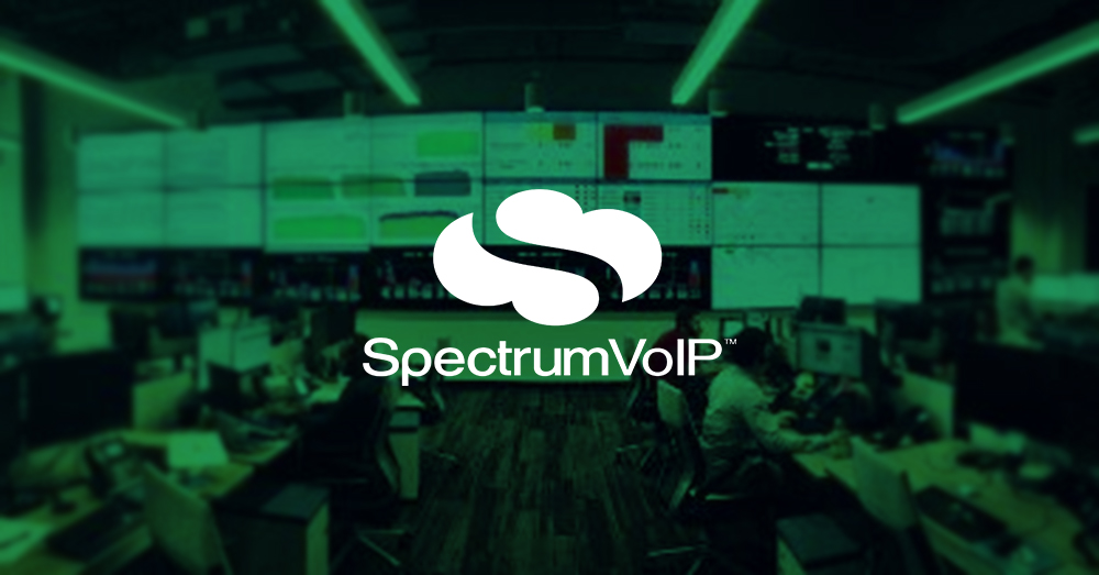 Empty SpectrumVoIP Network Operations Center with many workstations and a larger video wall displaying data dashboards with green overlay and logo