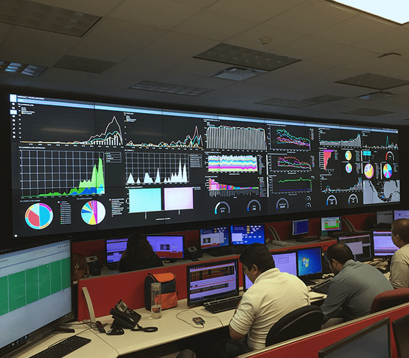 Employees working in a control room at workstations with video walls displaying data dashboards