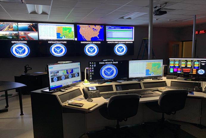 Empty EMA control room, with workstation, and a video wall displaying photography, data, and maps