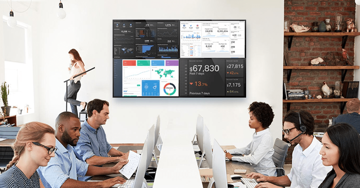 6 employees working at workstations in an office, with a video wall displaying data dashboards on the wall behind them