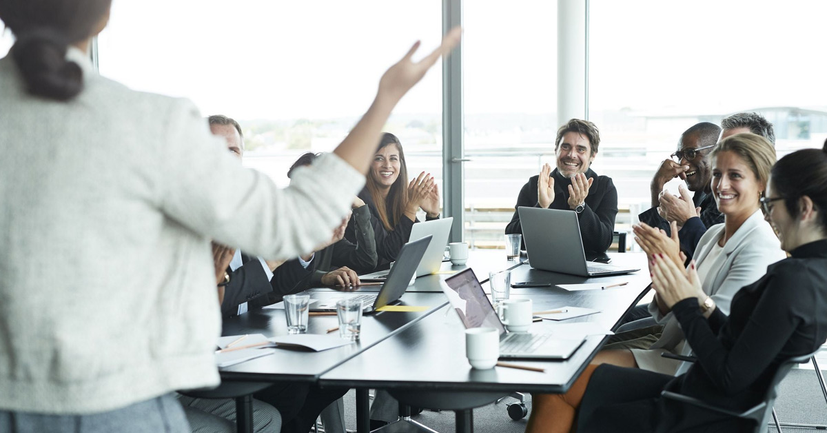 Employees in a meeting room clapping happily while a lady speaks to them