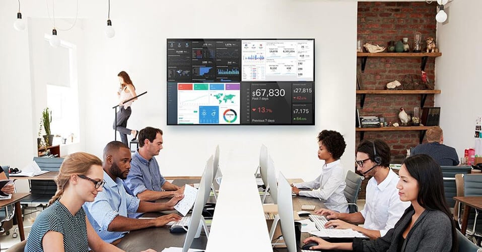 5 employees at their workstation, and a video wall displaying data dashboards