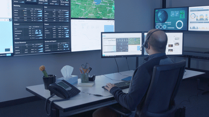 Logistics employee typing at his office desk in front of a video wall, then remotely from his home desk in front of a window and remotely