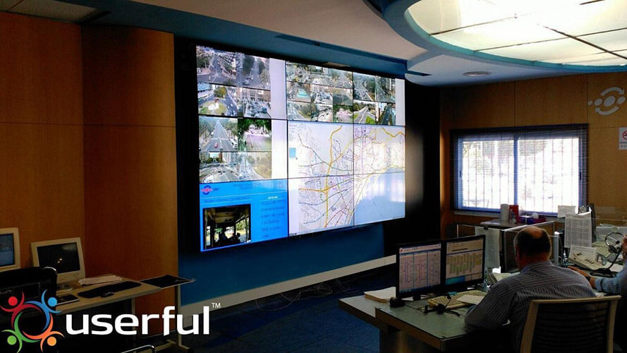 Two EMT employees monitoring transit operations through their workstations and a video wall displaying live camera footage, transit maps, and websites